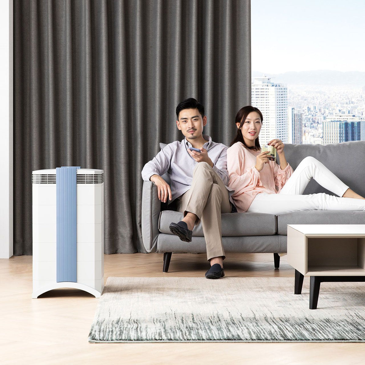 IQAir GC MultiGas XE Luftreiniger in Weiß neben Sofa, Paar entspannt im Wohnzimmer mit Skyline-Blick